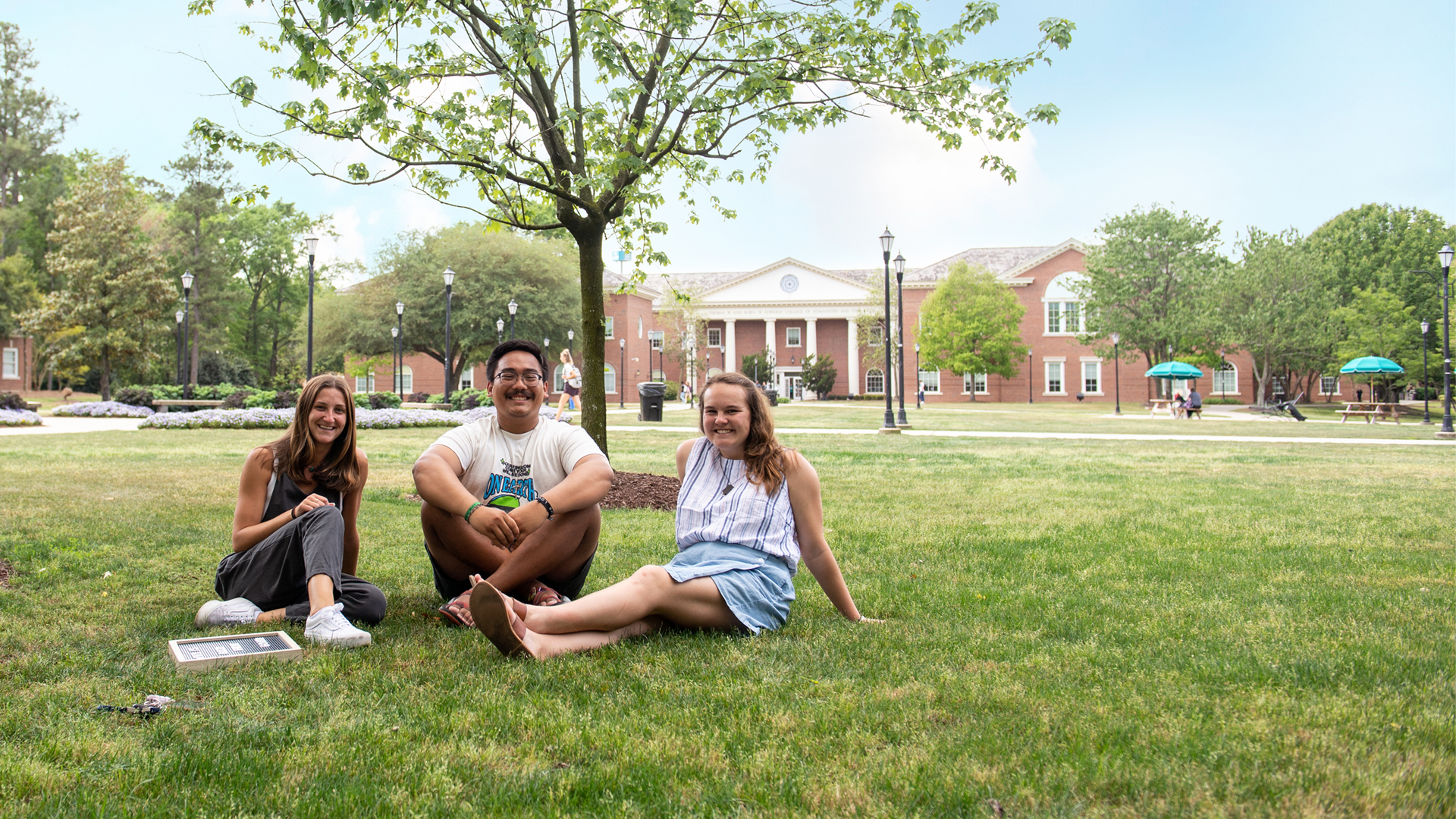 Three students site in the green grass on Prince Lawn, smiling while facing the camera with Edwards Building behind them.