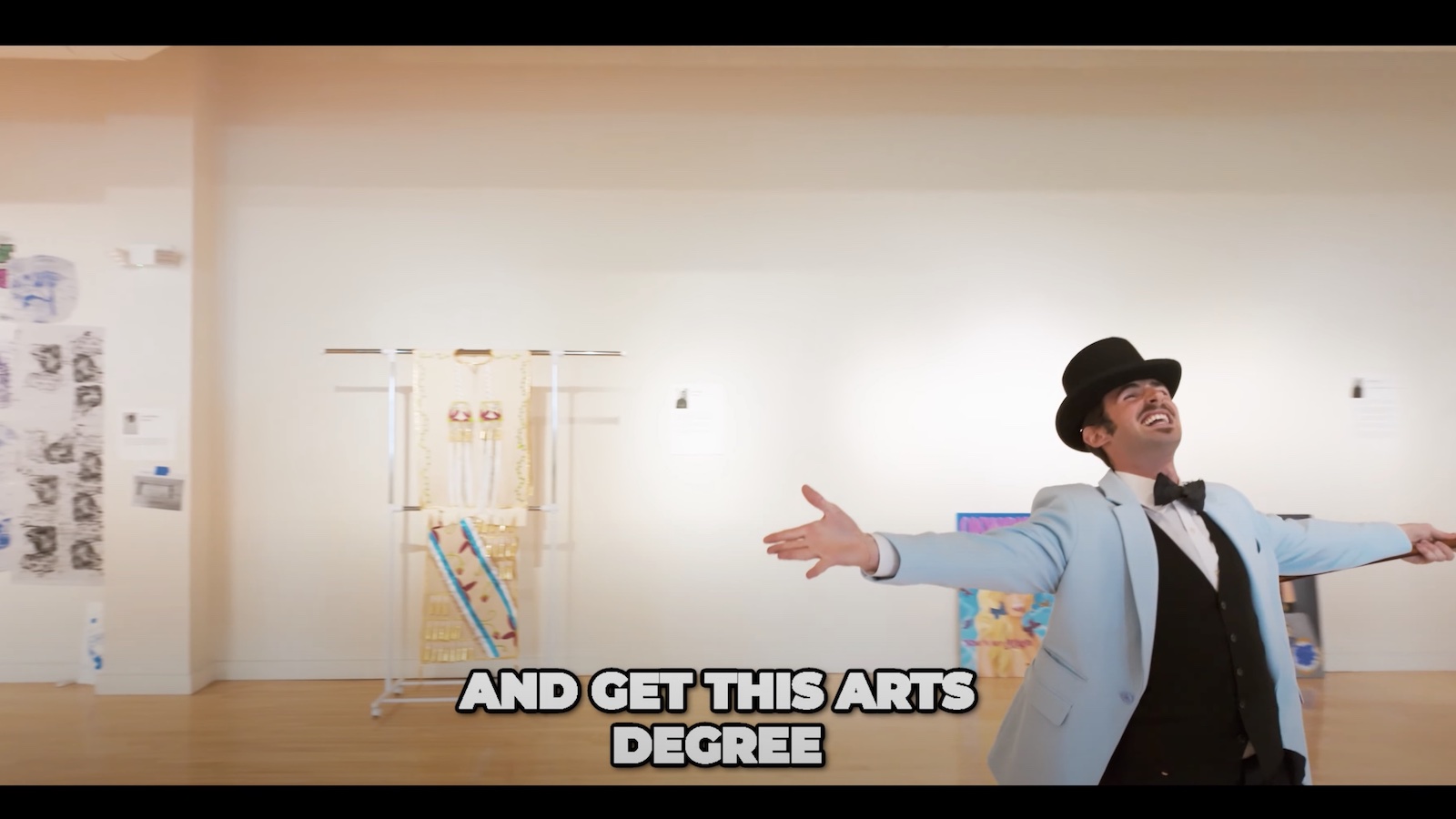 A student in a top hat and blue coat with bowtie dances around in-process art gallery space.