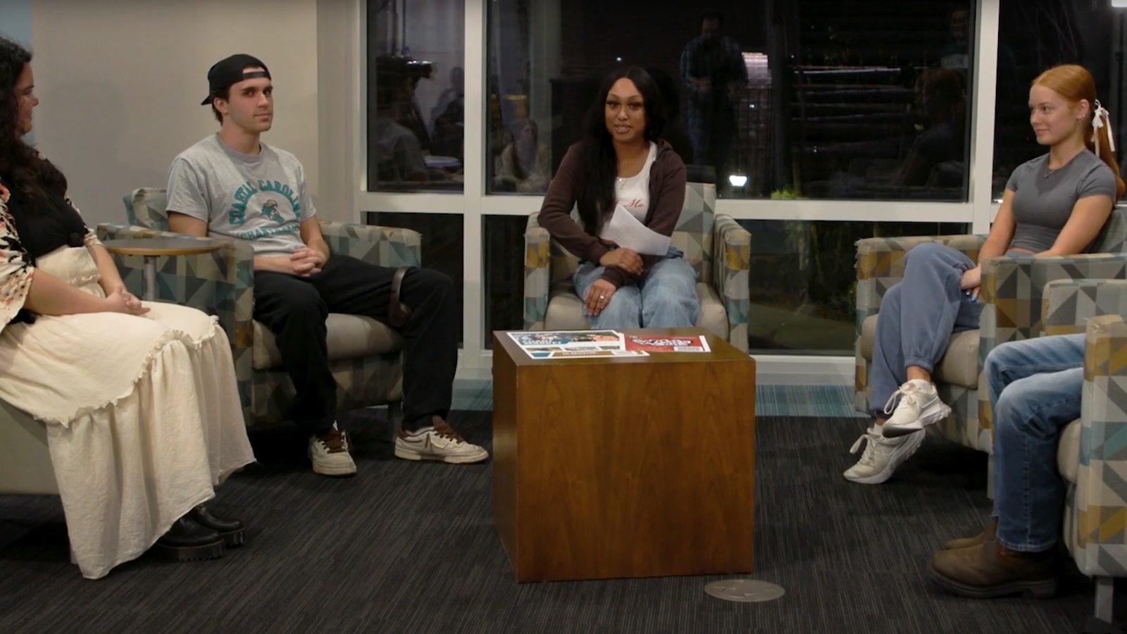 Four students (and a fifth offscreen) sit in teal chairs pulled into a semi-circle. Two are seniors, two are freshman, and one is a moderator.