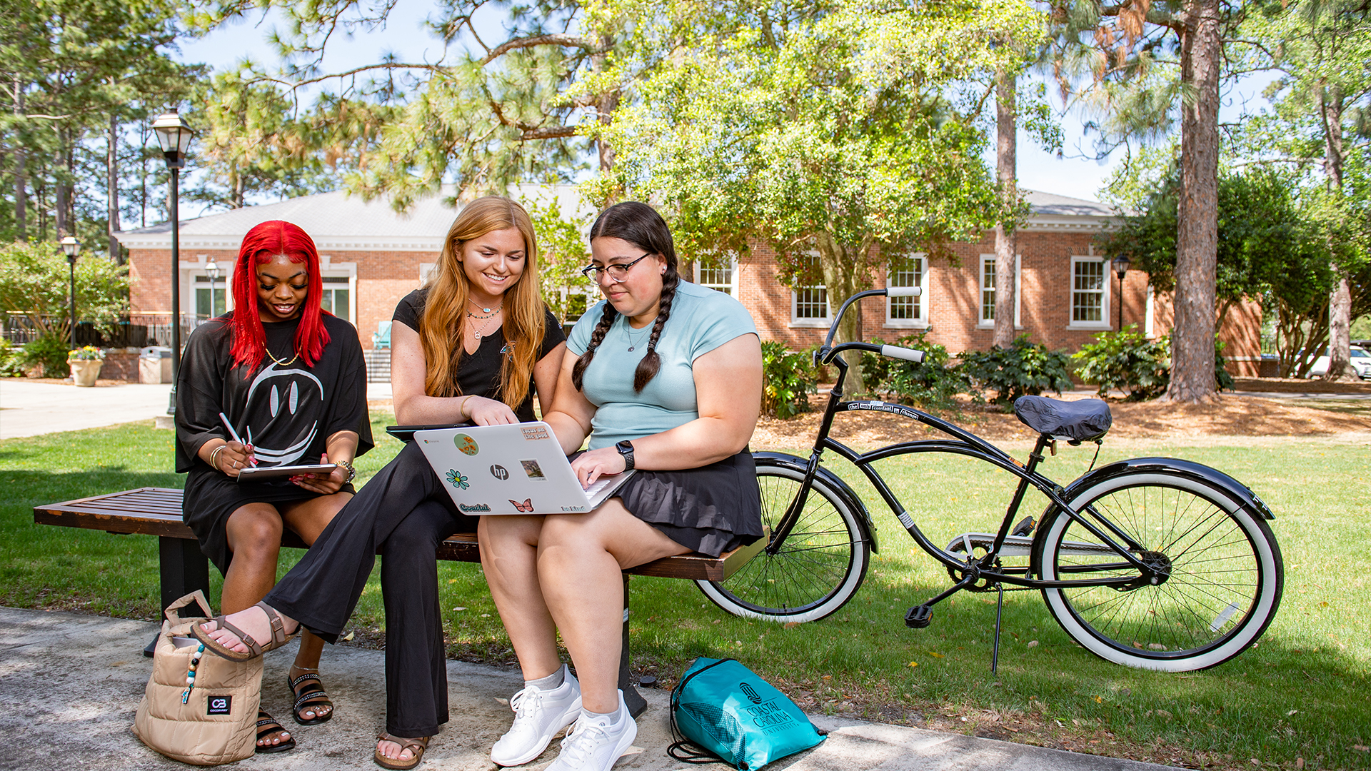 Three students sit on a bench, by a bicycle, at Blanton Park which is a central spot on campus for gathering and relaxing